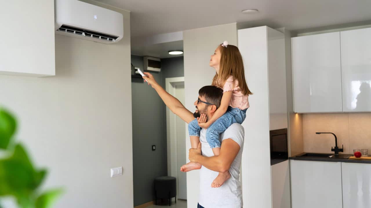 A man carries a girl on his shoulders as she uses a remote control to operate a wall-mounted air conditioner in their modern Alcoa, TN kitchen.