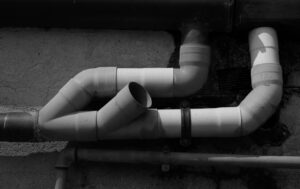 A network of white PVC pipes with multiple bends and joints is mounted against a rough exterior wall in black and white, reminiscent of industrial sites in Alcoa, TN.