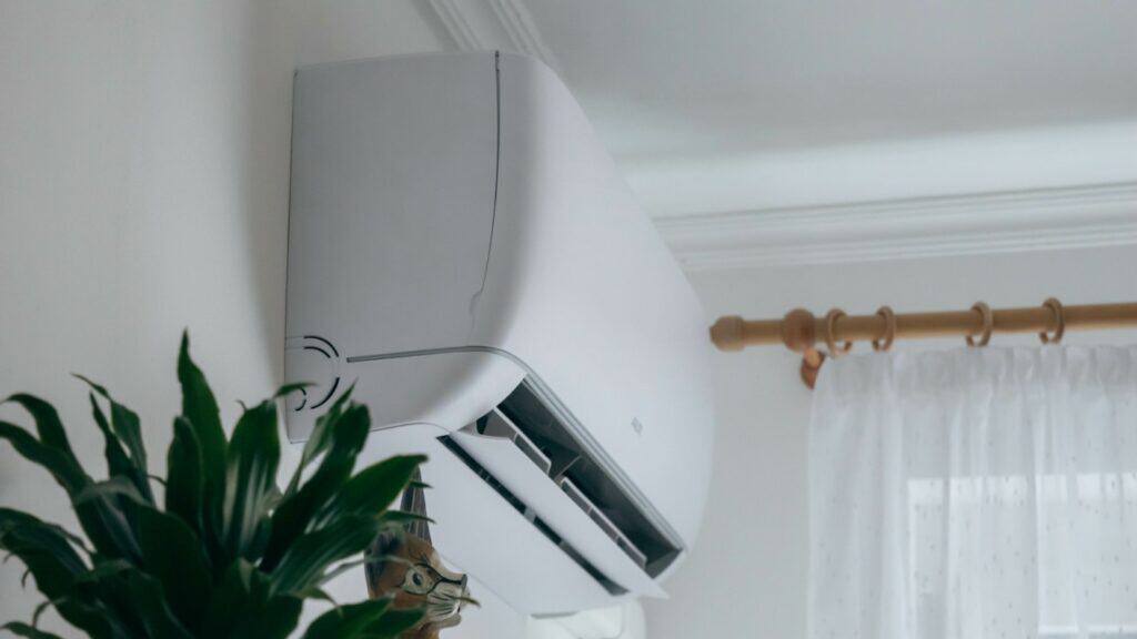 A wall-mounted air conditioner is installed above a window with a curtain rod and next to a green potted plant, creating a cool and inviting space in Alcoa, TN. Air conditioning replacement near me