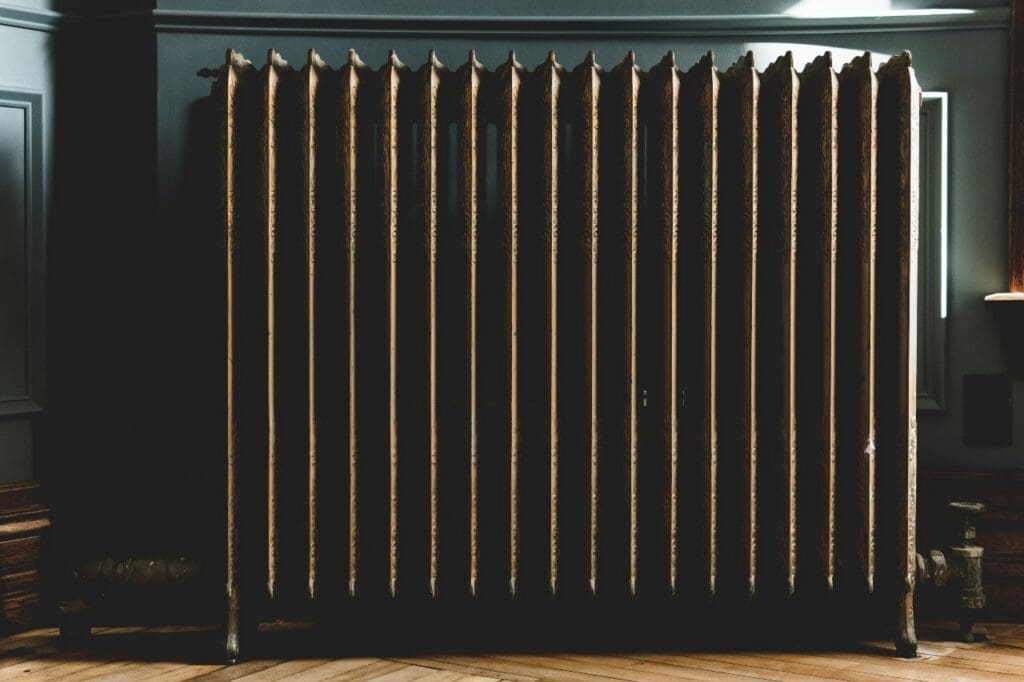 An old metal radiator with multiple vertical columns stands against a wall on a wooden floor in a dimly lit room in Alcoa, TN. Residential heating repair