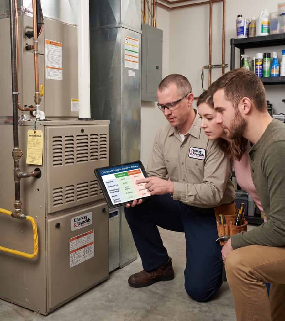 A technician shows a cost analysis chart on a tablet to two adults in a utility room next to a furnace and shelves with various household products.