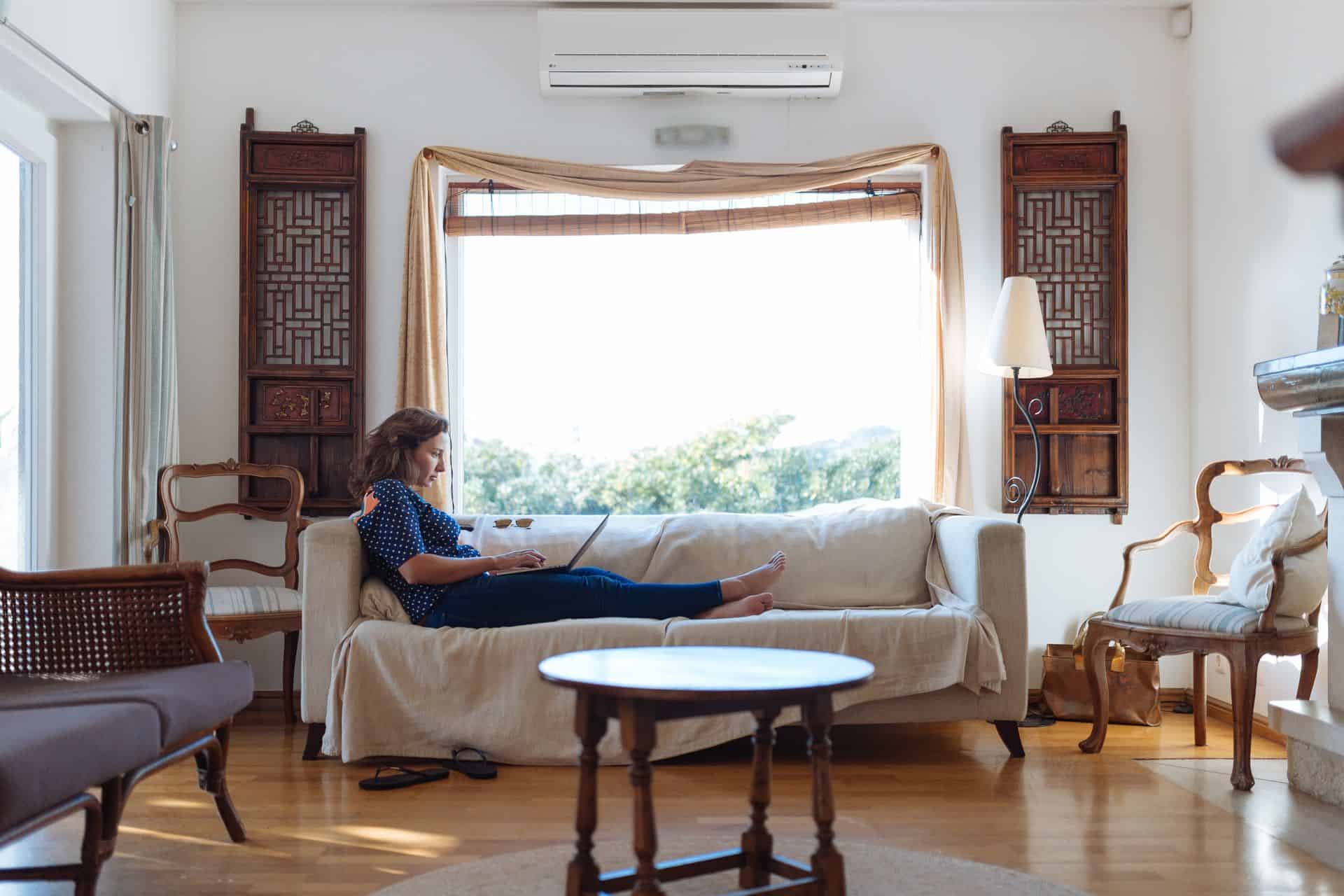 Woman sits on a beige sofa with a laptop in a bright living room with wooden furniture, large window, and sunlight streaming in.