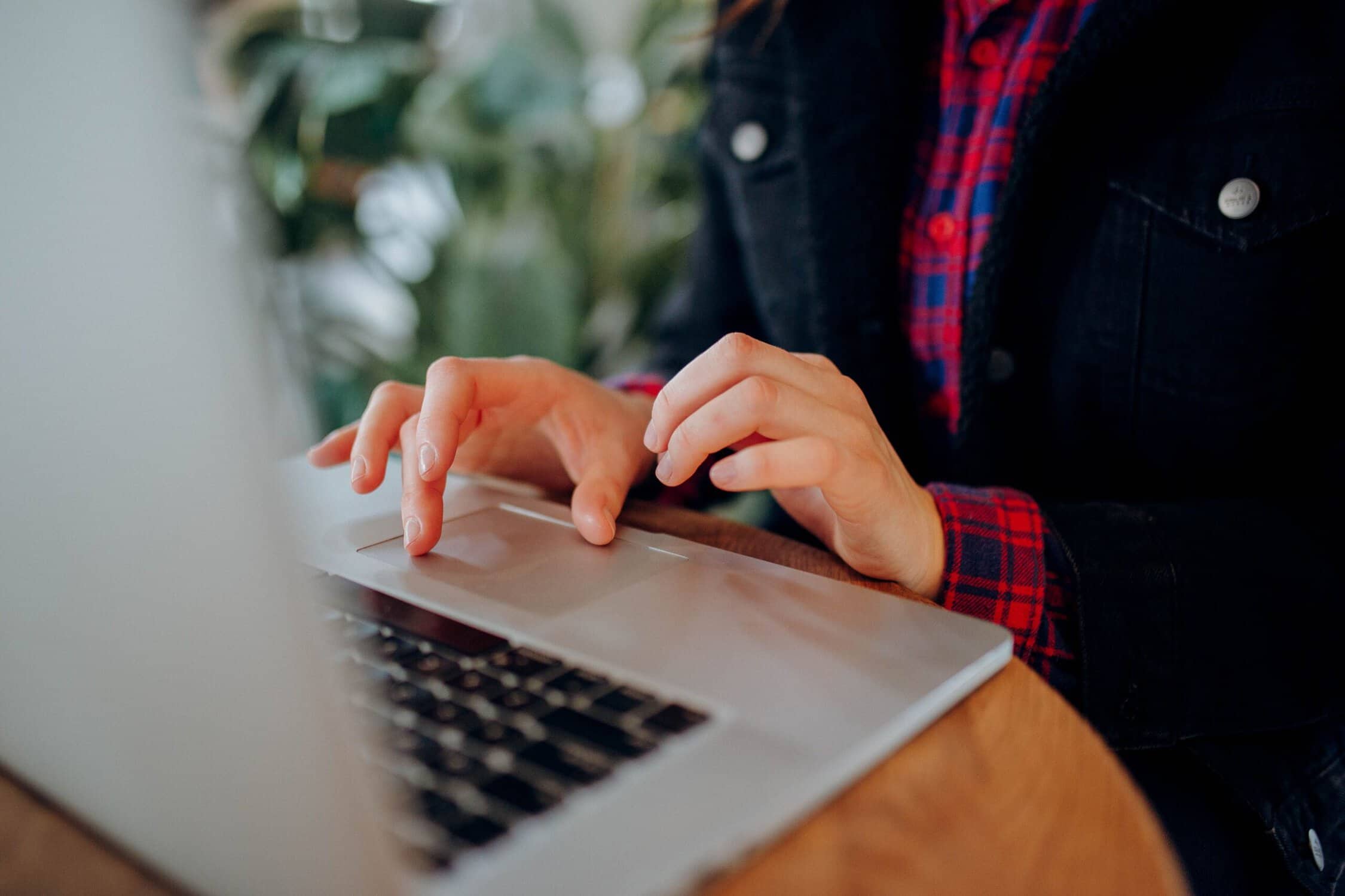 Person wearing a red plaid shirt and dark jacket typing on a laptop at a wooden table, with green plants blurred in the background.